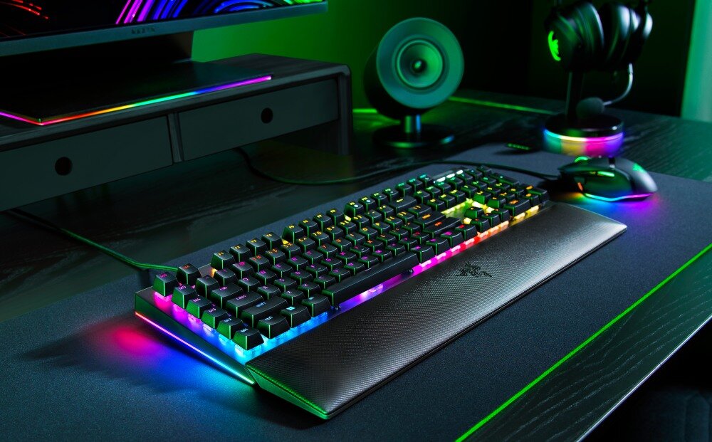 Na biurku znajduje się klawiatura z podświetleniem RGB, obok której leży myszka i słuchawki. W tle widoczny jest monitor oraz głośnik.