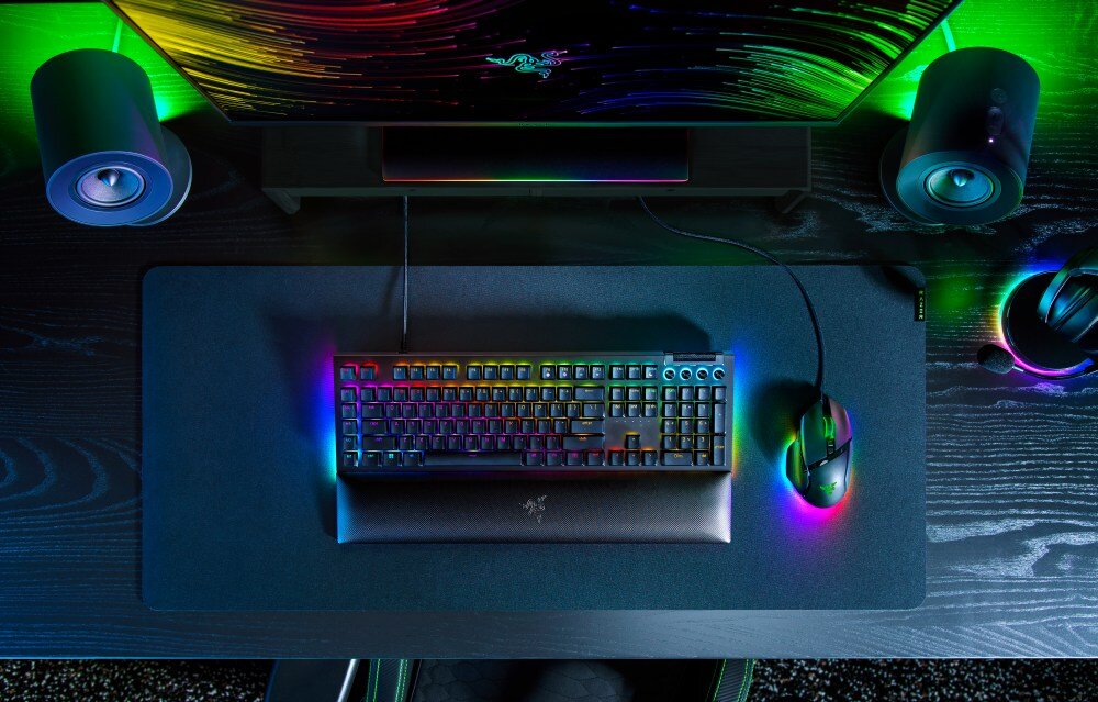 Na biurku znajduje się klawiatura z podświetleniem RGB oraz myszka, obok których widoczne są dwa głośniki i monitor. Logo producenta jest widoczne na klawiaturze i monitorze.