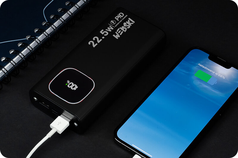 Na obrazie znajduje się czarny powerbank z napisem \'22.5W PD WEBSKI\' oraz wyświetlaczem pokazującym \'100%\'. Obok leży smartfon z ekranem pokazującym \'naładowano w 66%\'.
