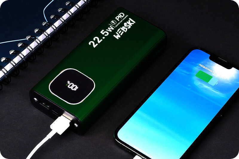 Zielony powerbank z wyświetlaczem pokazującym 100% oraz napisem \'22.5W PD WEBSKI\' leży obok smartfona z ikoną ładowania baterii. Obok znajduje się zeszyt w spiralnej oprawie.