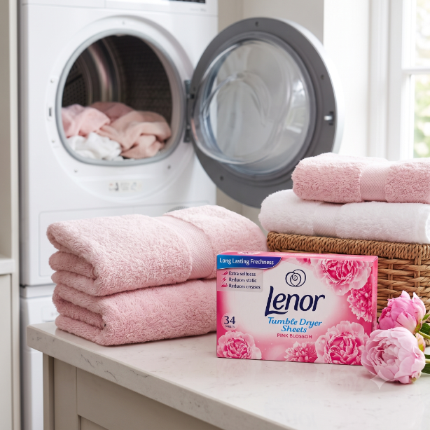 Na blacie znajduje się pudełko chusteczek do suszarki Lenor Tumble Dryer Sheets Pink Blossom 34 szt., obok którego leżą różowe ręczniki. W tle widać otwartą pralkę z różowym praniem.