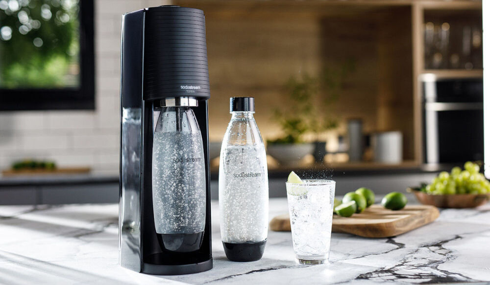 Na blacie kuchennym stoi czarny saturator z butelką wypełnioną gazowaną wodą oraz dodatkowa butelka i szklanka z napojem. Widoczne logo: sodastream.