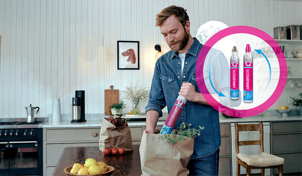 W kuchni na blacie stoi saturator, a na stole leżą cytryny i pomidory. W okręgu widoczne są trzy butelki z napisem \'SODASTREAM\'.