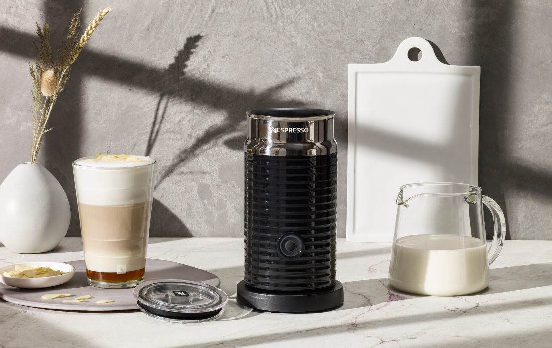 Na blacie znajduje się czarny spieniacz do mleka z napisem \'NESPRESSO\', obok szklanka z kawą, dzbanek z mlekiem i talerzyk z płatkami. W tle widać szarą ścianę i białą deskę do krojenia.