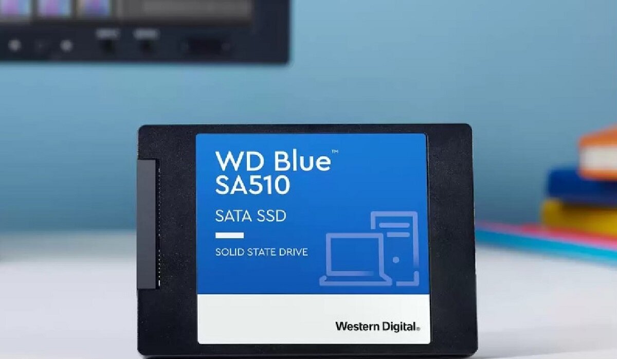 Na obrazie widoczny jest dysk SSD z etykietą \'WD Blue SA510 SATA SSD SOLID STATE DRIVE\' oraz logo \'Western Digital\'. W tle znajdują się rozmyte elementy, takie jak książki i ekran.
