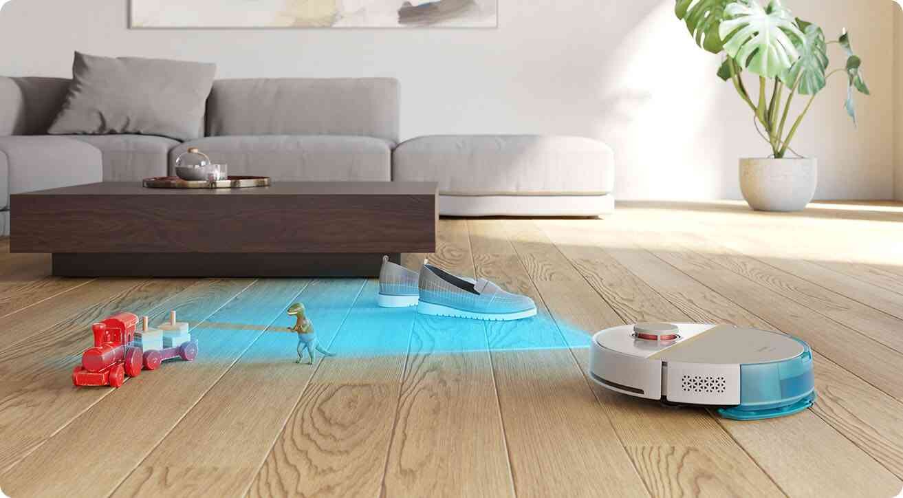 Robot sprzątający porusza się po drewnianej podłodze, omijając zabawki i buty. W tle widoczna jest szara sofa i stolik kawowy.