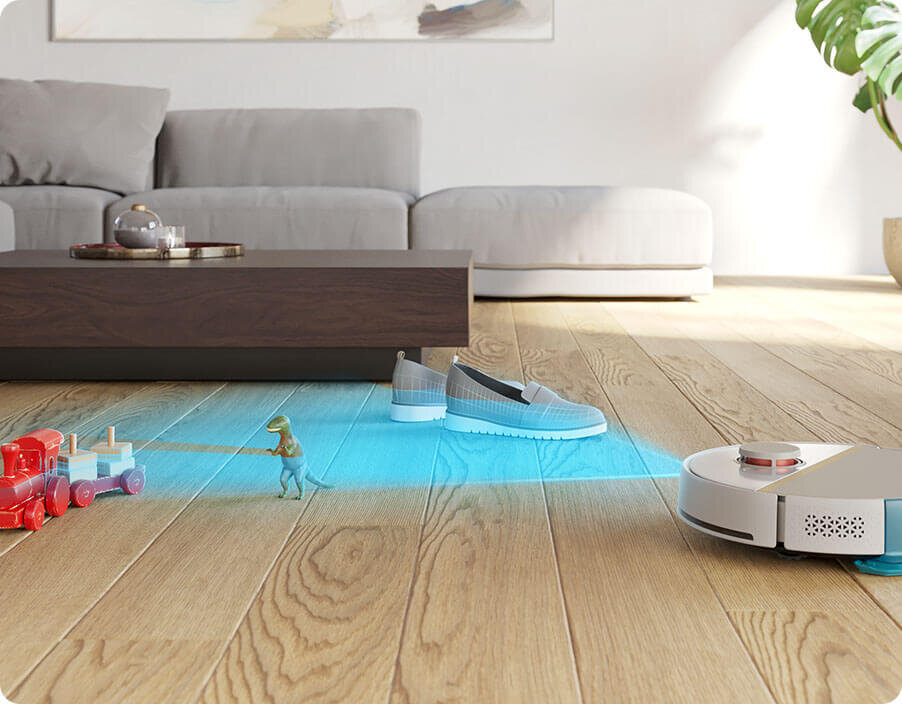 Robot sprzątający porusza się po drewnianej podłodze, omijając zabawki i buty. W tle widoczna jest szara sofa i niski stolik.