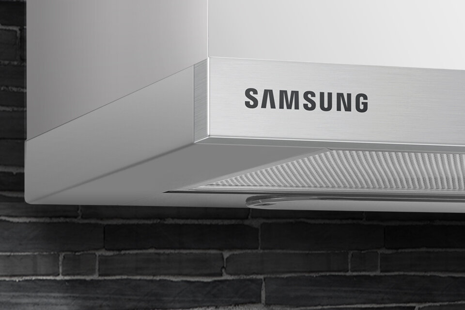 Samsung Minimalistyczny design. Wykończenie ze stali nierdzewnej Fragment okapu kuchennego z widocznym logo \'SAMSUNG\' na stalowej powierzchni. W tle ceglana ściana.