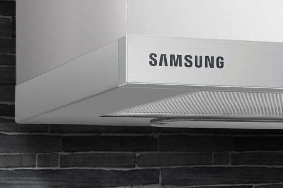 Samsung Minimalistyczny design. Wykończenie ze stali nierdzewnej Fragment okapu kuchennego z widocznym logo \'SAMSUNG\' na tle ciemnej, ceglanej ściany.