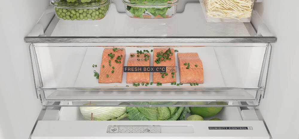 Wnętrze lodówki z widocznymi półkami, na których znajdują się pojemniki z warzywami i kawałki łososia z napisem \'FRESH BOX C°C\'. Na dolnej szufladzie widoczny jest napis \'HUMIDITY CONTROL\'.