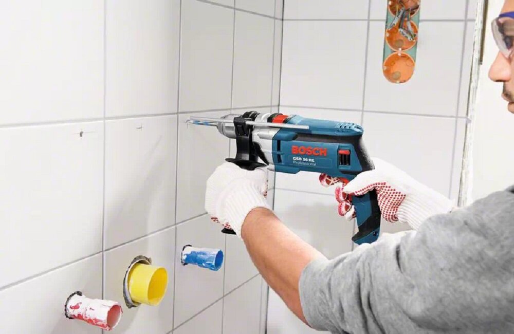 Osoba trzyma wiertarkę udarową BOSCH Professional GSB 16 RE, wiercąc otwór w białych płytkach ceramicznych. W tle widoczne są kolorowe rury wystające ze ściany.