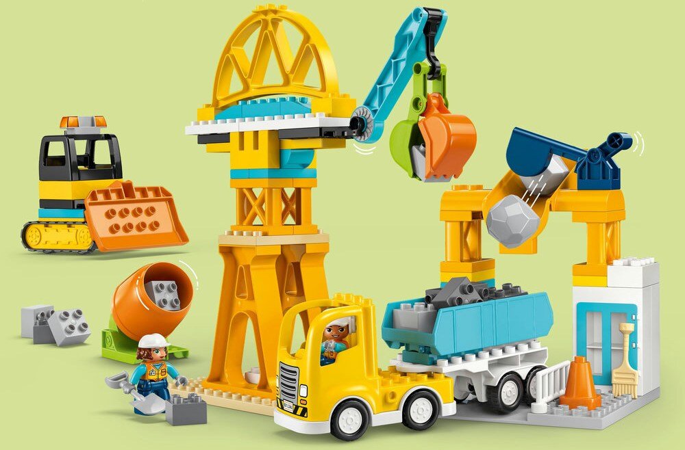 Na obrazku znajduje się zestaw klocków LEGO przedstawiający plac budowy z żółtym dźwigiem, ciężarówką i koparką. Widać także figurki pracowników budowy oraz elementy takie jak betoniarka i klocki imitujące kamienie.