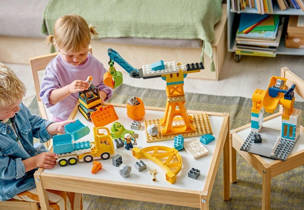 Na stole znajdują się klocki LEGO, w tym żółty dźwig, ciężarówka i inne elementy budowlane. Dwoje dzieci bawi się zestawem klocków.