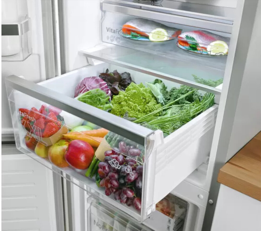 Otwarta lodówka z widocznymi szufladami wypełnionymi świeżymi warzywami i owocami, takimi jak sałata, winogrona, jabłka i marchewki. Na górnej półce znajdują się kawałki ryby z plasterkami cytryny, a na szufladzie widoczny jest napis \'VitaFresh\'.