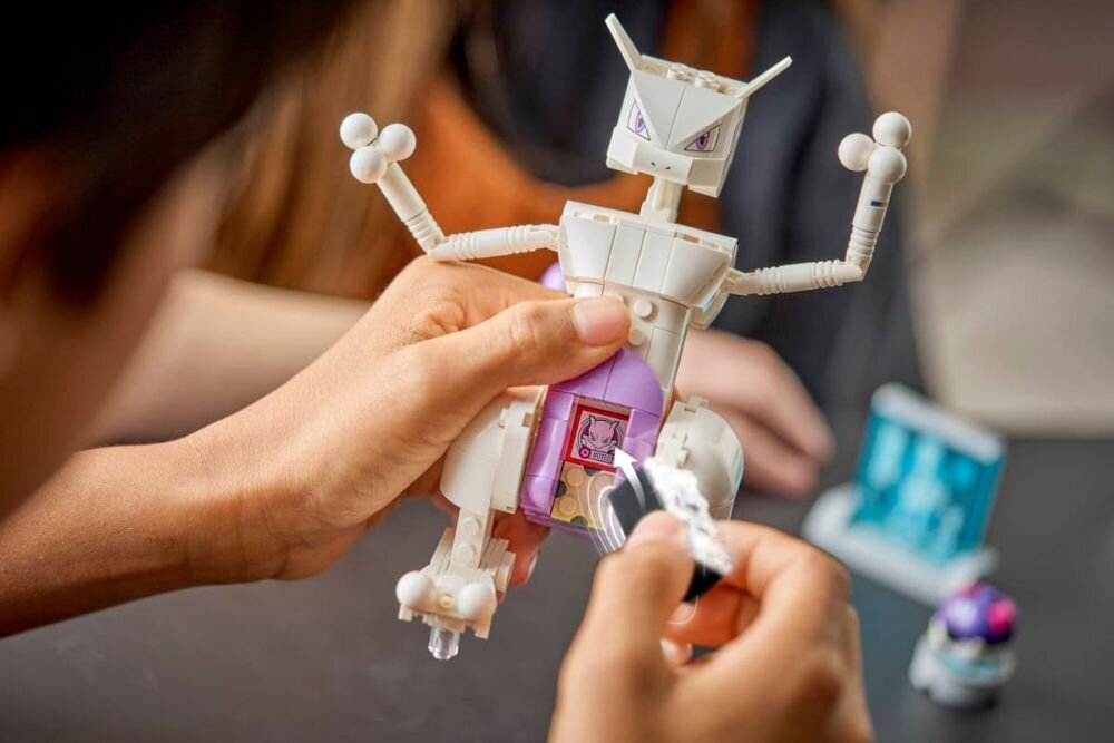 Osoba trzyma model z klocków przypominający robota z fioletowym elementem na środku. W tle widoczne są inne elementy zestawu LEGO.
