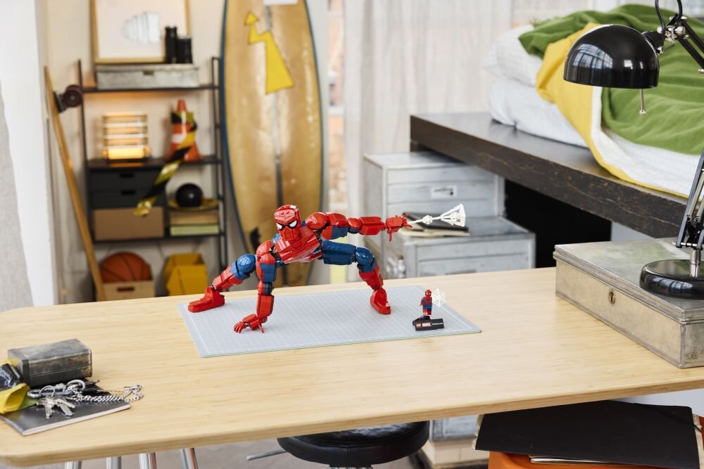 Na drewnianym biurku znajduje się duża figurka LEGO przedstawiająca Spider-Mana w dynamicznej pozie, obok której stoi mniejsza figurka na podstawce. W tle widać elementy wystroju wnętrza, takie jak lampa, łóżko i deska surfingowa.