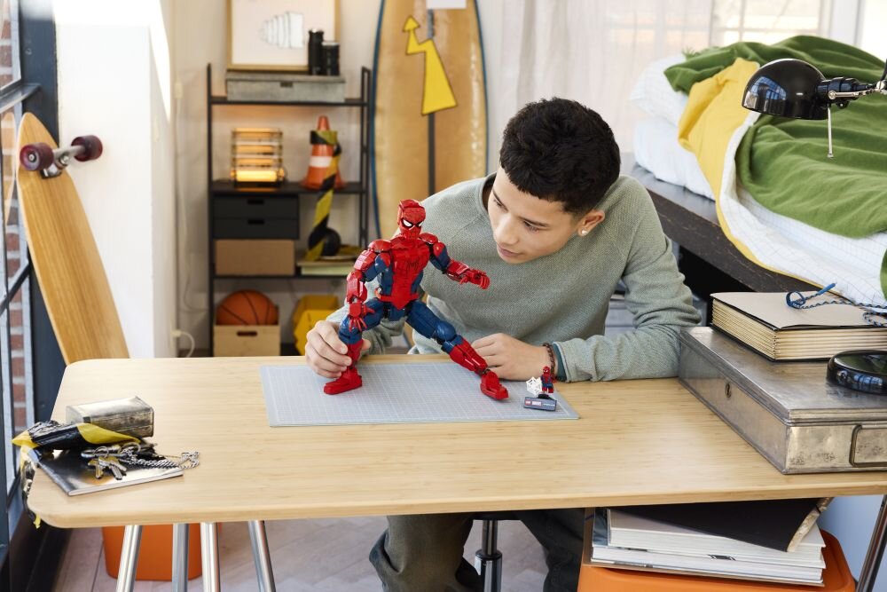 Na biurku znajduje się duża figurka Spider-Mana, którą trzyma osoba. W tle widać łóżko, deskorolkę i półkę z różnymi przedmiotami.