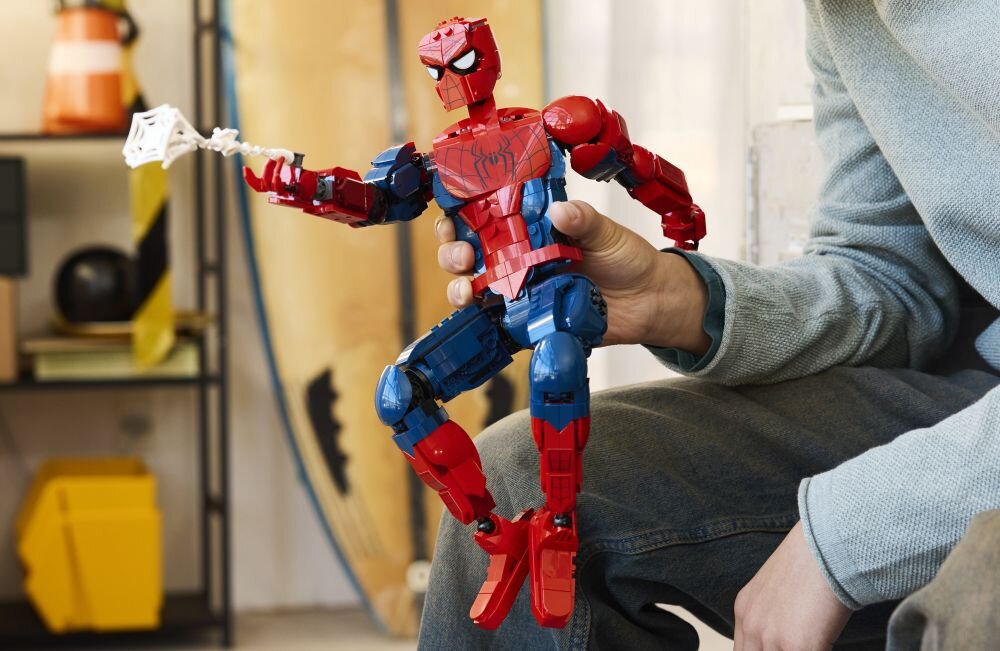 Osoba trzyma dużą figurkę Spider-Mana zbudowaną z klocków LEGO, w kolorach czerwonym i niebieskim, z pajęczyną w ręce. W tle widoczne są elementy wnętrza, takie jak deska surfingowa i półka.