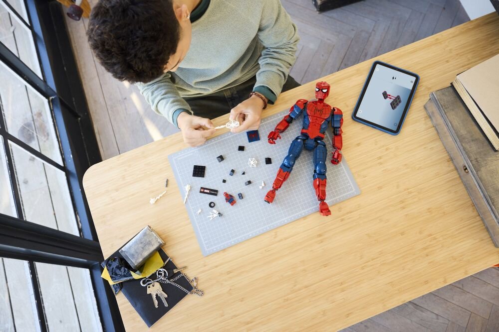 Osoba składa zestaw LEGO z figurką Spider-Mana na drewnianym stole, obok leży tablet z instrukcją. Na macie znajdują się różne elementy LEGO, a obok leży złożona figurka Spider-Mana.