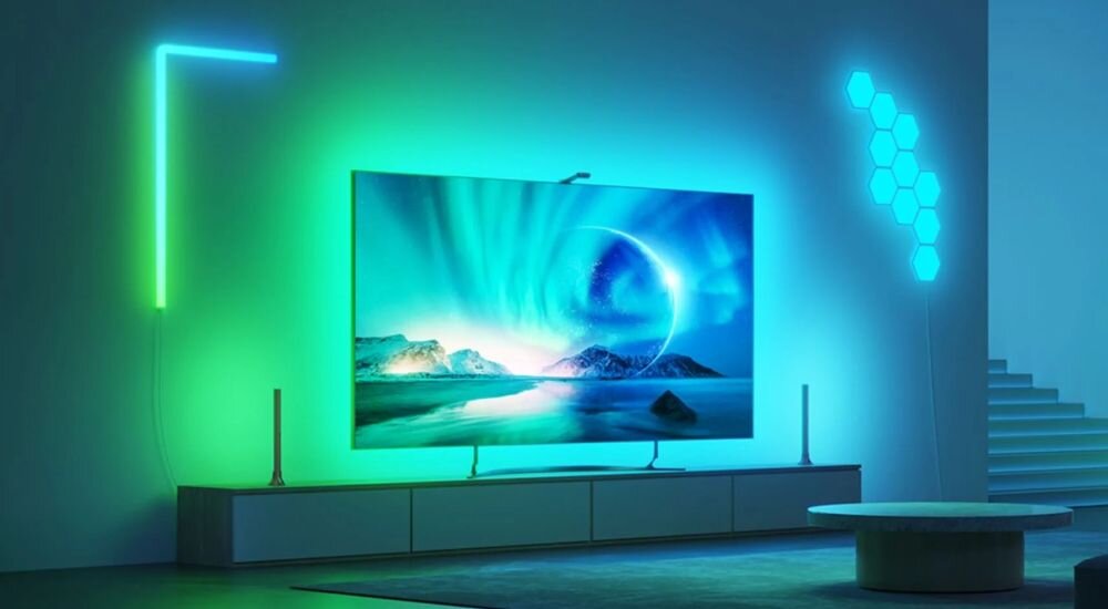 Pokój z dużym telewizorem wyświetlającym krajobraz z zorzą polarną, otoczony kolorowym oświetleniem LED. Na ścianie widoczne są również sześciokątne panele świetlne.