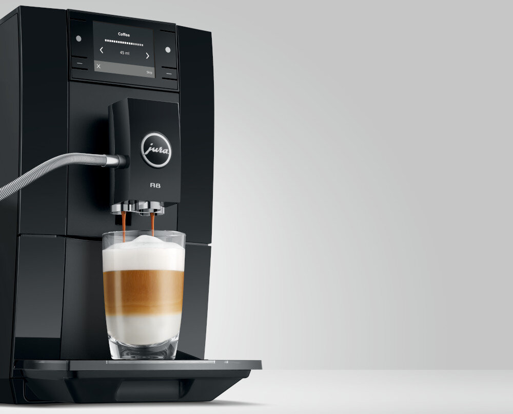 Czarny ekspres do kawy z logo \'jura\' i oznaczeniem \'A8\' przygotowuje kawę z mleczną pianką w szklance. Na wyświetlaczu widoczny jest napis \'Coffee\' oraz ustawienia dotyczące ilości wody i mocy kawy.