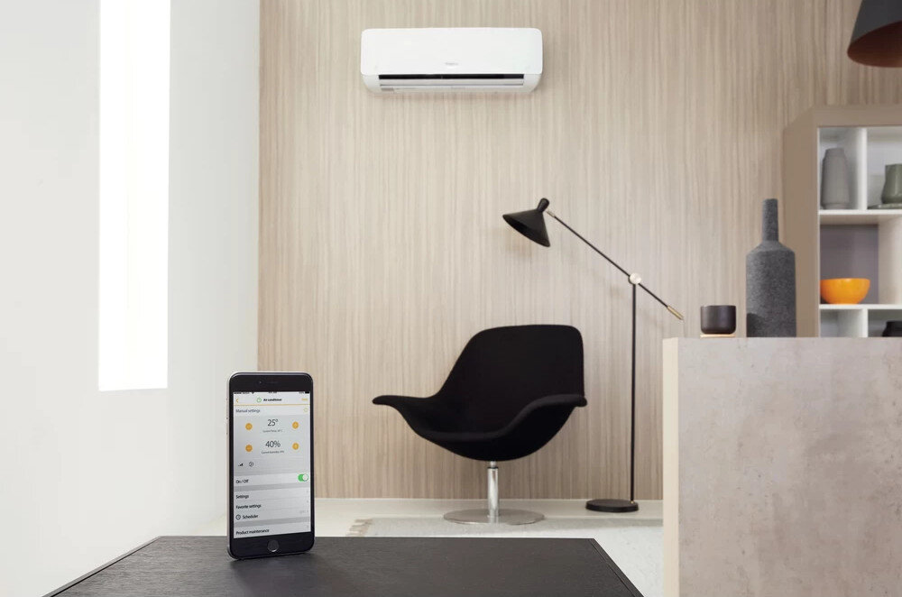 W nowoczesnym wnętrzu znajduje się klimatyzator zamontowany na ścianie oraz czarny fotel i lampa stojąca. Na pierwszym planie widoczny jest smartfon z aplikacją do sterowania klimatyzatorem, na ekranie widnieje temperatura 25°C i wilgotność 40%.