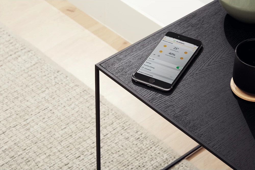 Na czarnym stoliku leży smartfon z wyświetlonym interfejsem aplikacji do sterowania klimatyzatorem. Obok znajduje się czarny kubek na drewnianej podstawce.