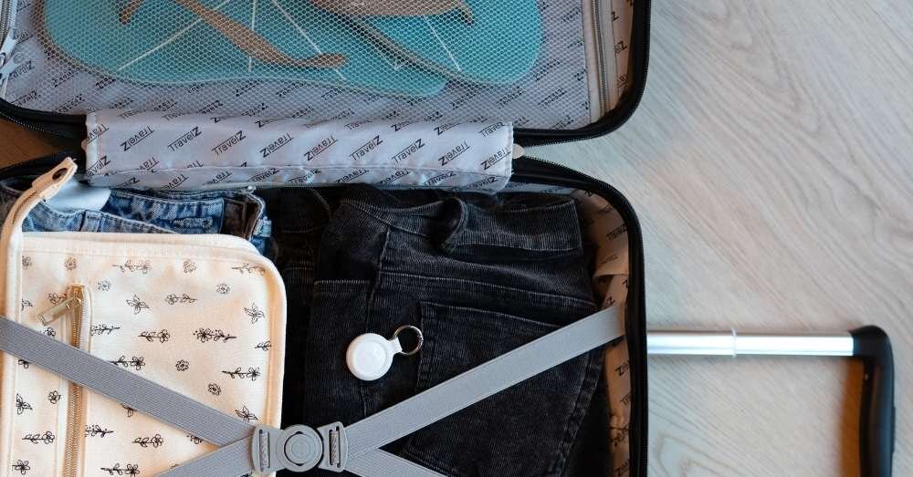 Otwarta walizka z ubraniami, w tym jeansami i torbą z wzorem, oraz białym lokalizatorem przypiętym do paska. W tle widoczne są klapki i materiał z napisem \'ZWEAR\'.