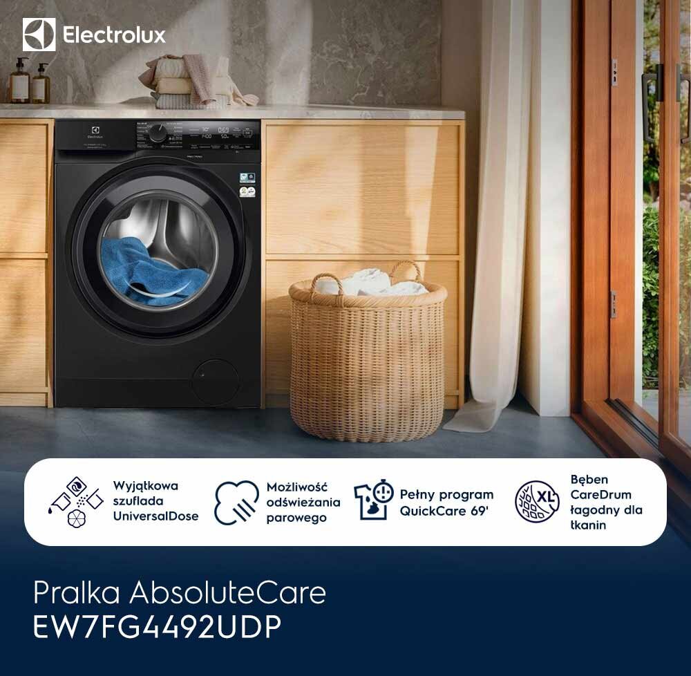 Na obrazie widoczna jest czarna pralka Electrolux z otwartym bębnem, w którym znajduje się niebieskie pranie. Obok pralki stoi kosz wiklinowy z białym praniem, a w tle widać drewniane szafki i duże okno.