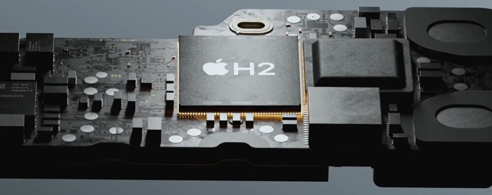 Obraz przedstawia płytkę drukowaną z widocznym chipem oznaczonym logo Apple i napisem \'H2\'. Wokół chipa znajdują się różne elementy elektroniczne, takie jak kondensatory i złącza.