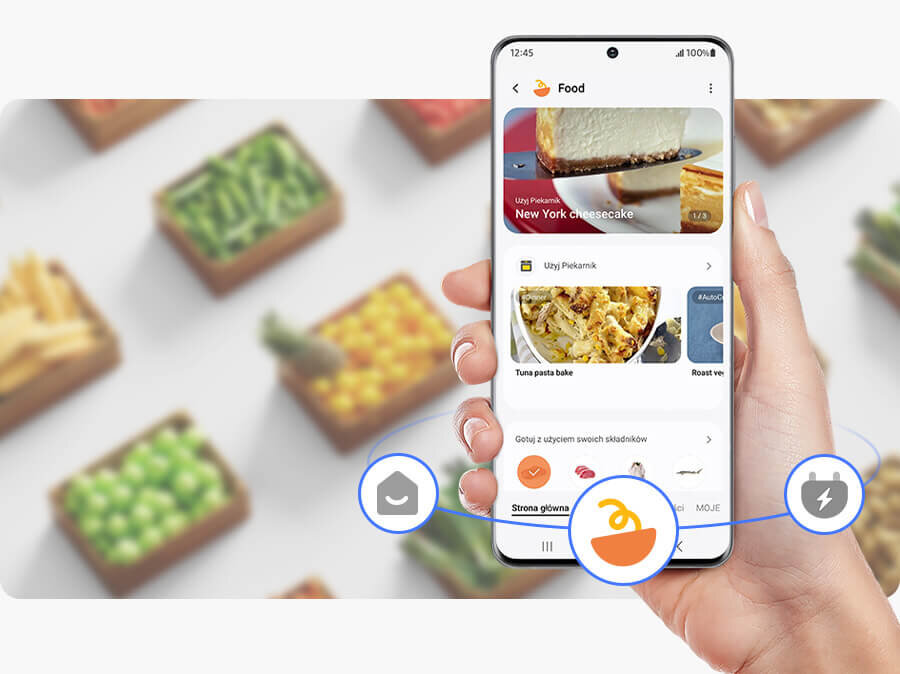 wyszukaj przepisy w aplikacji SmartThings, a potrzebne składniki dodaj do listy zakupów Osoba trzyma smartfon z aplikacją kulinarną, na ekranie widoczny jest przepis na \'New York cheesecake\'. W tle rozmyte pudełka z warzywami.