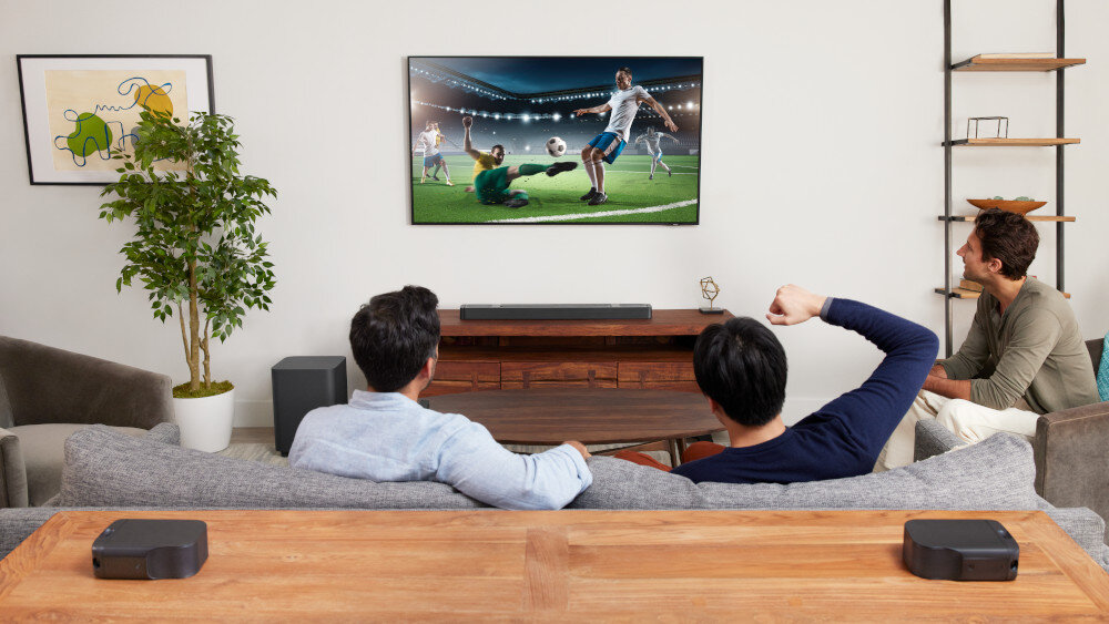 Trzech mężczyzn siedzi na kanapie w salonie, oglądając mecz piłki nożnej na dużym telewizorze. Na szafce pod telewizorem znajduje się soundbar, a w pomieszczeniu widoczne są elementy dekoracyjne, takie jak roślina i obraz na ścianie.