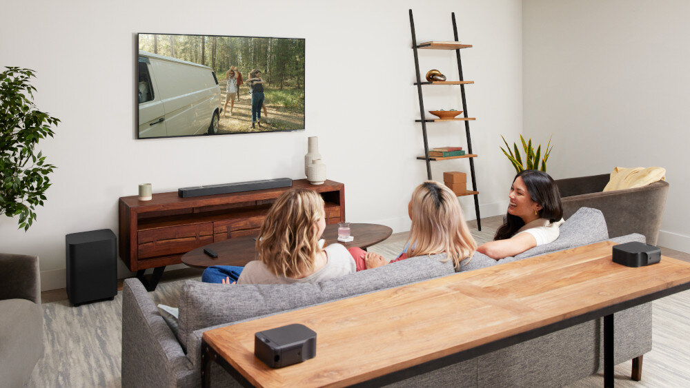 W salonie na ścianie wisi telewizor, pod którym znajduje się soundbar na drewnianej szafce. Trzy osoby siedzą na kanapie, oglądając film, a obok szafki stoi subwoofer.