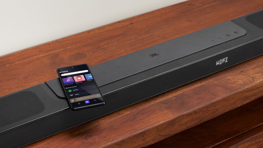 Na drewnianej powierzchni znajduje się czarny soundbar z logo JBL i napisem \'WiFi\'. Na soundbarze leży smartfon z wyświetlonym interfejsem aplikacji muzycznej.