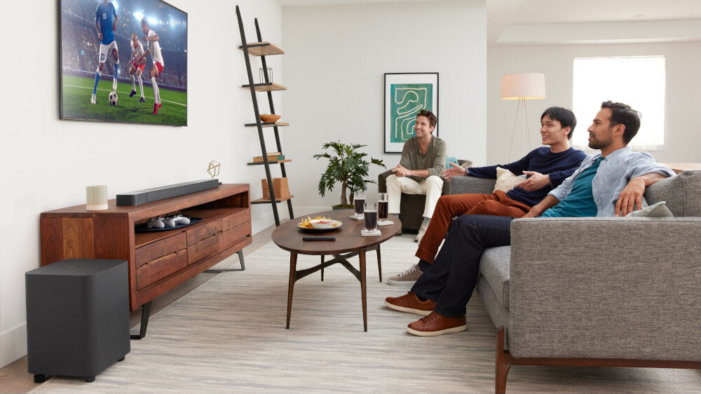 W salonie na szafce stoi soundbar, a obok niego znajduje się subwoofer. Na ścianie wisi telewizor, na którym wyświetlany jest mecz piłki nożnej.