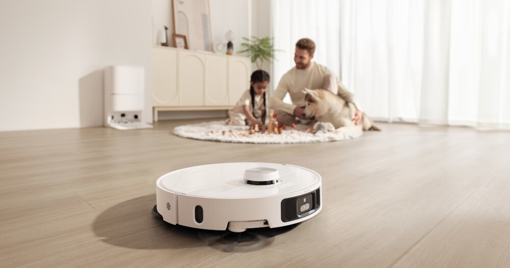 Na podłodze znajduje się biały robot sprzątający, a w tle widać osobę z dzieckiem i psem siedzących na dywanie.