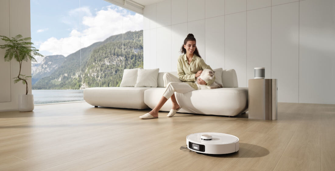 Na drewnianej podłodze znajduje się biały robot sprzątający. W tle widać osobę siedzącą na kanapie z kotem oraz duże okno z widokiem na góry i jezioro.