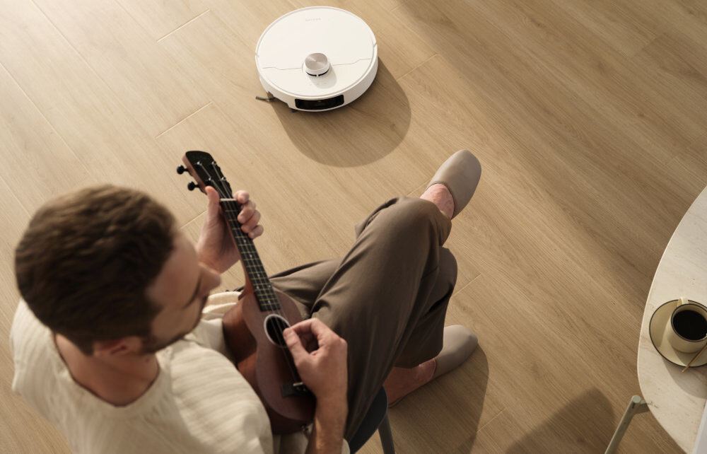 Na drewnianej podłodze znajduje się biały robot sprzątający. Obok siedzi osoba grająca na ukulele, a na stoliku stoi filiżanka z napojem.