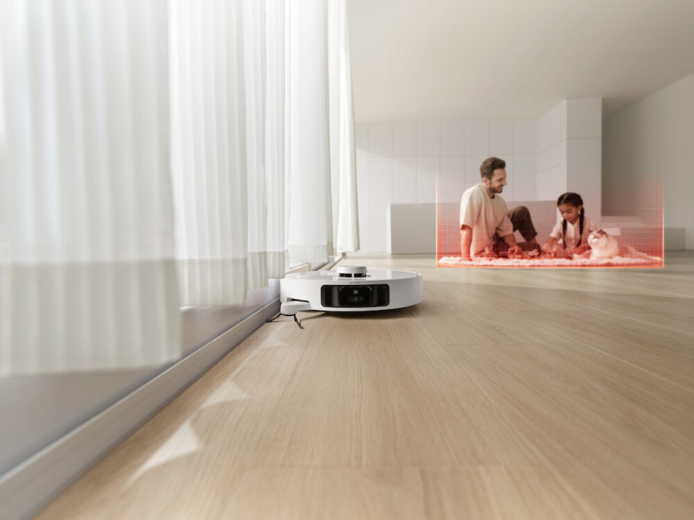 Na drewnianej podłodze znajduje się biały robot sprzątający. W tle widać dwie osoby siedzące na dywanie z kotem, otoczone czerwonym polem.