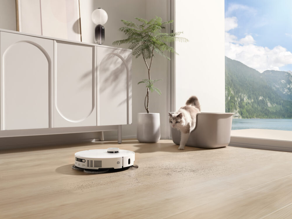 Robot sprzątający porusza się po drewnianej podłodze, obok niego znajduje się kot wychodzący z kuwety. W tle widać jasną szafkę, roślinę w doniczce i duże okno z widokiem na góry i jezioro.