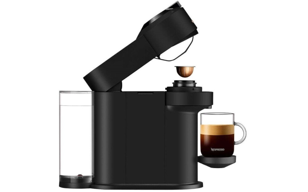 Czarny ekspres do kawy z otwartą pokrywą, w której znajduje się kapsułka. Obok ekspresu stoi szklanka z kawą, a na niej widnieje napis \'Nespresso\'.