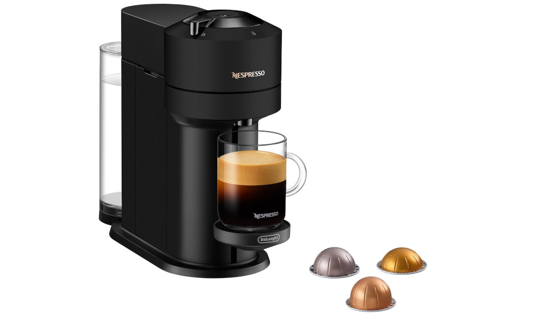 Czarny ekspres do kawy z logo Nespresso i Delonghi, z przezroczystym pojemnikiem na wodę oraz filiżanką z kawą. Obok ekspresu znajdują się trzy kapsułki w różnych kolorach.