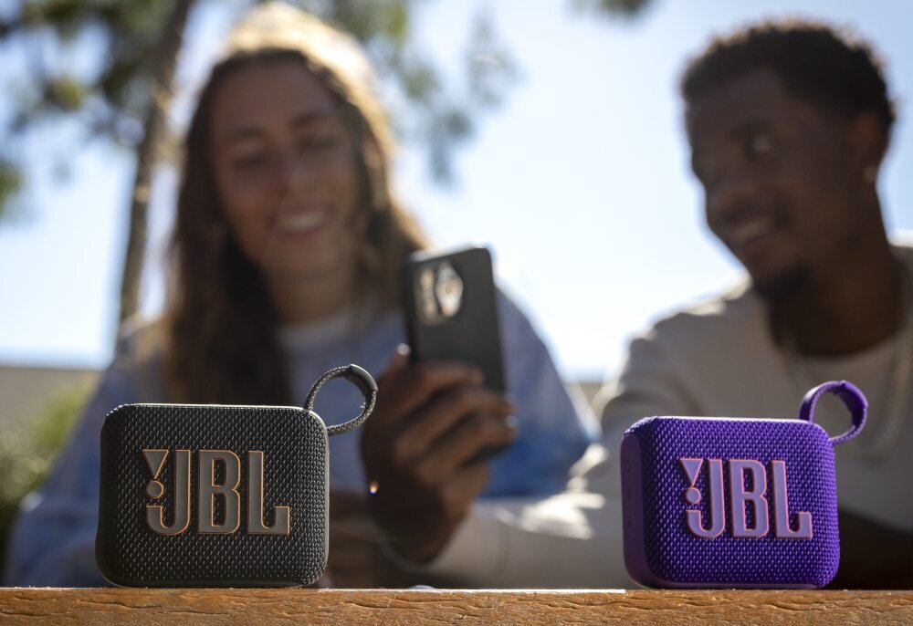 Na pierwszym planie znajdują się dwa głośniki mobilne z logo JBL, jeden czarny, drugi fioletowy. W tle widoczne są dwie osoby trzymające telefon.