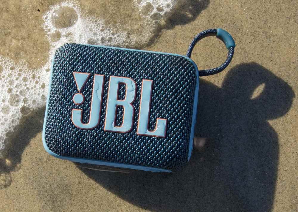 Na piasku leży niebieski głośnik z logo \'JBL\', obok którego widać wodę z pianą.