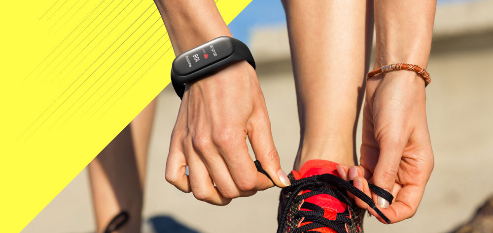 Osoba wiąże sznurowadła w sportowym bucie, mając na nadgarstku czarny smartband z wyświetlaczem pokazującym czas i kroki. Tło jest rozmyte, a po lewej stronie znajduje się żółty pasek z ukośnymi liniami.