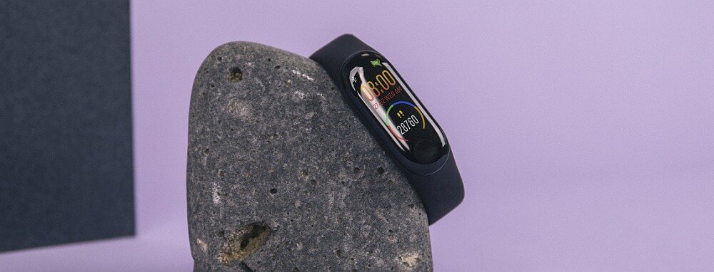 Czarny smartband z kolorowym wyświetlaczem pokazującym godzinę 08:00 i liczbę kroków 8760, umieszczony na szarej, kamiennej powierzchni na fioletowym tle.