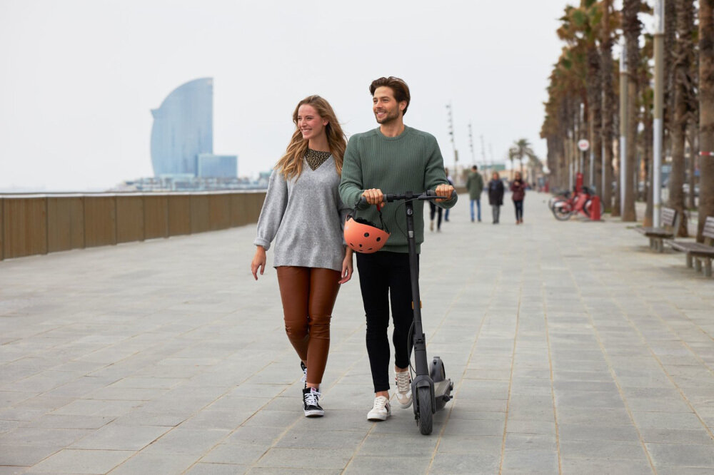 Dwie osoby spacerują obok hulajnogi elektrycznej na promenadzie z palmami. W tle widoczny jest nowoczesny budynek o charakterystycznym kształcie.