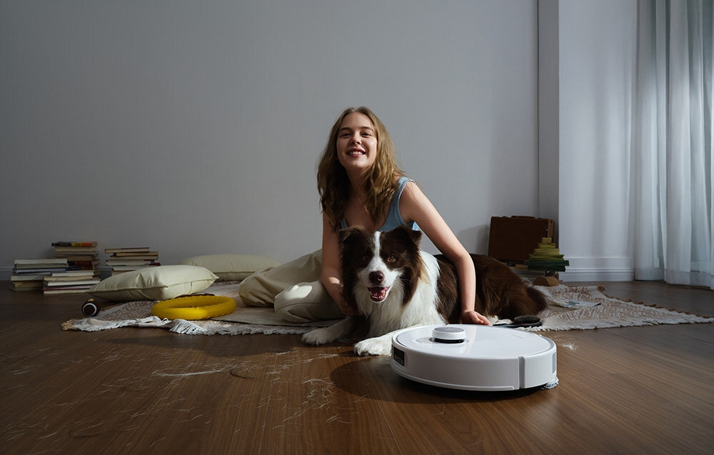 Na drewnianej podłodze znajduje się biały robot sprzątający. Obok niego siedzi osoba z psem, a w tle widać poduszki i stosy książek.