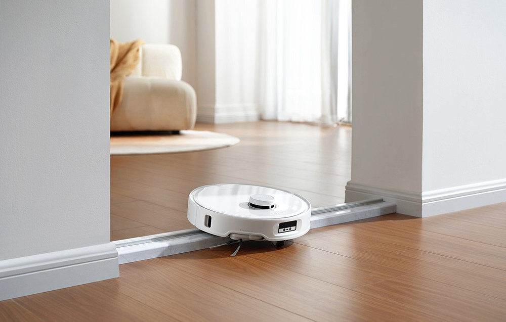 Biały robot sprzątający porusza się po drewnianej podłodze, przekraczając metalowy próg. W tle widoczna jest część jasnej kanapy i duże okno.