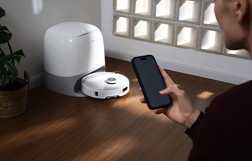 Na drewnianej podłodze znajduje się biały robot sprzątający obok stacji dokującej. Osoba trzyma w ręku smartfon, patrząc na robota.